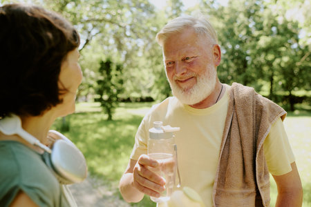 Gray-haired man holding bottle of water and smiling to his brunette wife after run in parkの写真素材