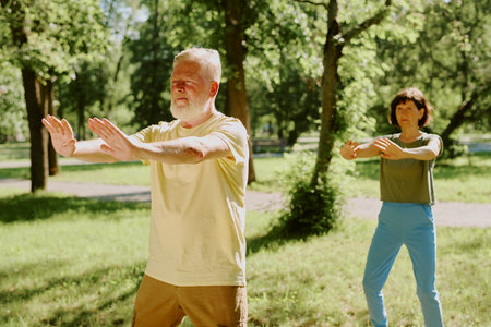 Senior silver-haired man with thick beard and brunette woman dressed in sportswear holding qigong pose to improve breath during open air workout in parkの写真素材