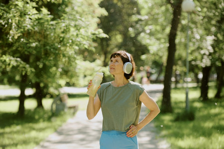 Senior woman drinking pure water during small break after running, she enjoying music in her headphonesの写真素材