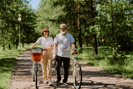 Senior loving couple dressed in casual clothes and wearing sunglasses having bike walk in park during warm summer dayの写真素材