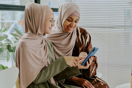 Two Muslim women smiling while looking at smartphone, surrounded by plants in modern interior, bonding and sharing moments together, both wearing hijabs and stylish clothingの写真素材
