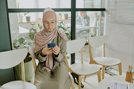 Muslim woman in hijab focusing on smartphone while sitting in modern office environment with plants and natural light entering through large windowsの写真素材