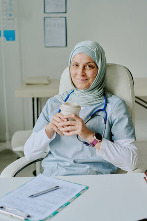 Medical professional in hijab sitting in clinic smiling while holding coffee cup, with documents on desk and medical charts on wall in backgroundの写真素材
