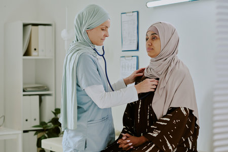 Medical professional wearing scrubs and hijab checking patients vitals in modern healthcare setting, both women wearing hijabs, standing near medical equipmentの写真素材
