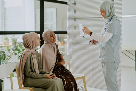 Doctor engaging with two Muslim women in medical office, holding document, providing an explanation and advice regarding health care options, women dressed in modest attire in seated positionの写真素材