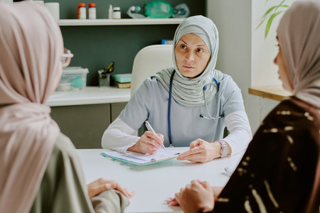Doctor wearing hijab sitting in office while consulting patients and taking notes. Medical instruments and supplies visible in background creating professional atmosphereの写真素材