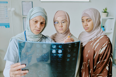 Medical professional analyzing X-ray while two patients wearing hijabs attentively observing the results in a clinic settingの写真素材
