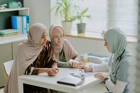 Three females discussing medical care in an office setting with medical items on desk, doctor in white coat listening attentively during consultationの写真素材