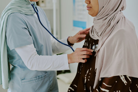 Doctor examining patient with stethoscope during medical check-up in clinical setting. Patient wearing hijab and doctor in scrubs, focusing on chest areaの写真素材