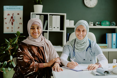 Two women sitting at a medical office, with one wearing a stethoscope and the other in a patterned hijab, posing for the camera and smilingの写真素材