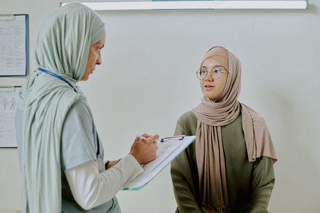 Healthcare professional in headscarf talking to patient while taking notes. Medical consultation focusing on understanding and discussing patients health needsの写真素材