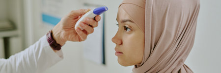 Profile view of woman wearing hijab receiving temperature check using infrared thermometer device in medical setting without natural lightの写真素材
