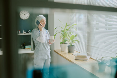 Nurse wearing scrubs and headscarf standing by window, speaking on phone while holding coffee cup, plants and window blinds in background, medical office settingの写真素材