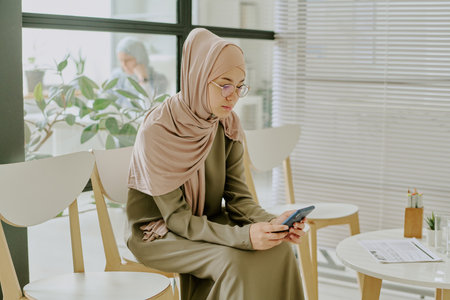Middle Eastern woman wearing hijab and green dress sitting on white chair using smartphone in modern office setting with plant and large windowsの写真素材