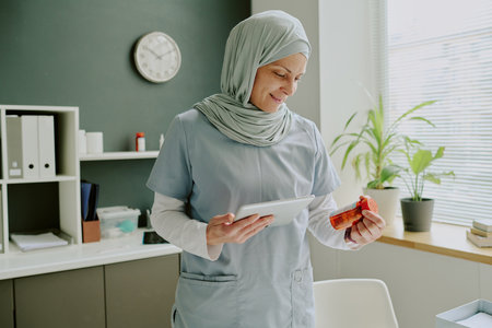 Smiling nurse wearing hijab standing in medical office holding digital tablet and red prescription bottle, surrounded by organized shelves, plants, and office suppliesの写真素材