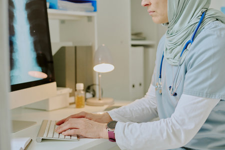 Female doctor wearing hijab working at office desk, engaging with computer and medical equipment, showing dedication to her professionの写真素材