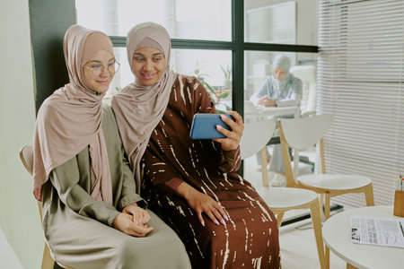 Women wearing traditional attire seated together, sharing and viewing content on smartphone in a cozy room with natural light from large windows. Background shows someone working at deskの写真素材