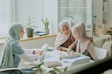 Female healthcare professional wearing hijab consulting two young women in modern medical office. Various medical tools and plants are visible on white deskの写真素材