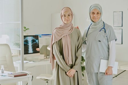 Portrait of two female healthcare professionals standing in modern medical office, one in hijab and other in medical uniform, looking directly at camera, smilingの写真素材