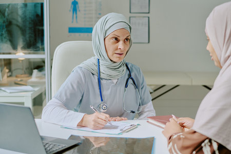 Medical professional wearing hijab sitting at desk, engaging with patient during consultation, with stethoscope around her neck and medical charts in backgroundの写真素材