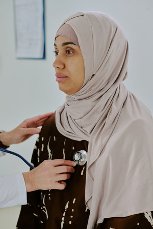 Medical professional using stethoscope for examining patient in clinic setting Patient wearing hijab, showing trust and comfort during examinationの写真素材