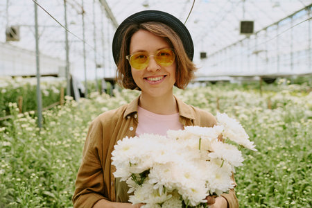 Woman holding white flowers while standing in greenhouse wearing yellow sunglasses and black hat delighting in serene environment of lush greeneryの写真素材