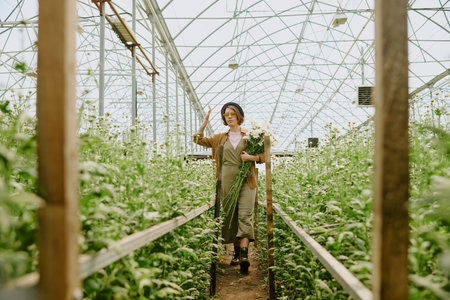 Woman admiring flowers in greenhouse with natural light filtering through roof, creating serene ambiance. She holds a bouquet, surrounded by rows of thriving plantsの写真素材