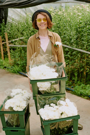 Smiling woman arranging bouquets of white flowers in a greenhouse, wearing sunglasses and a hat, surrounded by greenery and flower arrangementsの写真素材