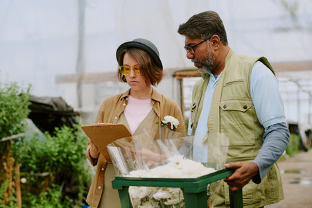 Two people discussing flower arrangements in greenhouse, standing next to lush greenery and flowers, one holding clipboard, another holding flower bouquetの写真素材