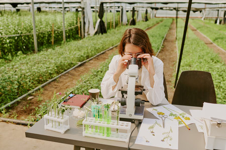 Scientist sitting in laboratory surrounded by plant samples and equipment observing specimens through microscope in greenhouse environmentの写真素材