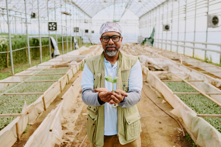 Portrait of elderly man smiling while holding a small seedling in spacious greenhouse with lush plants growing in backgroundの写真素材