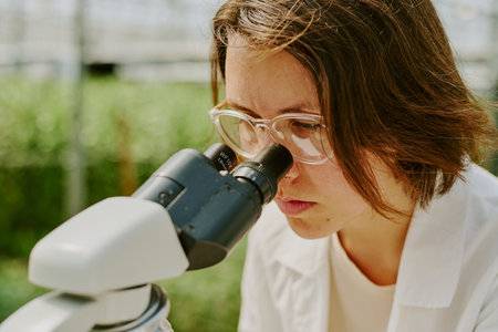 Scientist intently observing sample through microscope in laboratory setting wearing white lab coat and safety glasses, background blurred with laboratory equipmentの写真素材