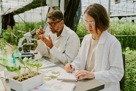 Two scientists working in a lab examining plant specimens surrounded by lush greenery. Researching plant growth under controlled conditions in a greenhouse settingの写真素材
