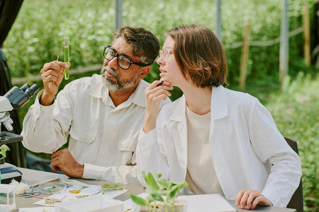 Two scientists analyzing plant samples outdoors, holding test tube and discussing results, surrounded by laboratory equipment and greenery under a shade structureの写真素材