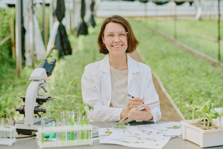 Scientist analyzing plants in laboratory greenhouse setting surrounded by equipment and samples, smiling and sitting at desk with clear test tubes, microscope, and green plantsの写真素材