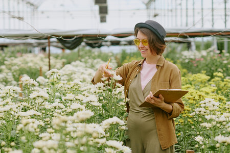 Happy woman wearing hat and sunglasses while inspecting flowers in greenhouse and taking notes. Surrounded by blooming plants and clear sky visible through glass roofの写真素材