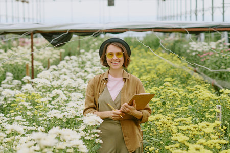 Smiling woman standing among blooming flowers in outdoor garden, holding notebook and wearing hat. Glasses adding stylish touch against green and floral backdropの写真素材