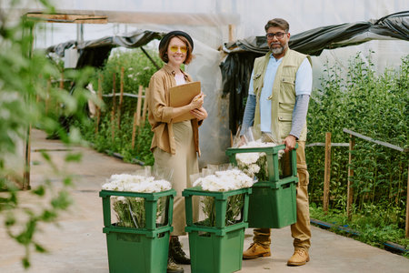 Farmers standing in greenhouse, holding flowers and clipboard while smiling, showcasing freshly harvested blooms in green crates, and wearing protective clothingの写真素材