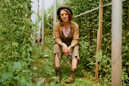 Woman sitting on a chair in garden surrounded by lush greenery wearing yellow sunglasses and boots holding hand tool showing relaxed and thoughtful expressionの写真素材