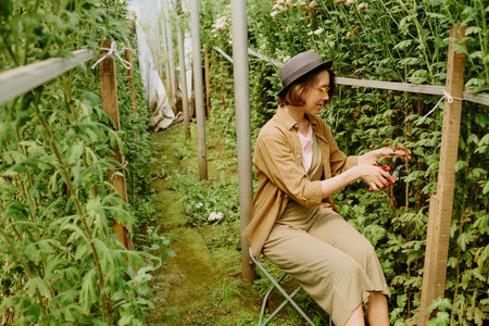 Woman wearing hat carefully pruning plants in greenhouse. Focused on maintaining healthy foliage in an enclosed, lush environmentの写真素材