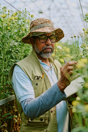 Elderly man wearing beige hat and glasses, surrounded by blooming yellow plants in greenhouse, taking notes or making observations in small notepad, concentrating deeplyの写真素材