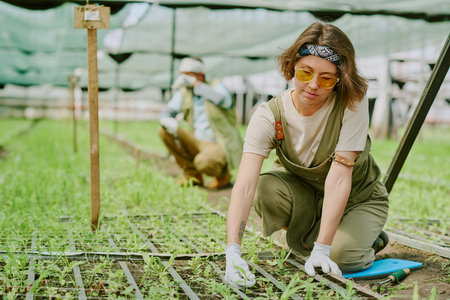 Woman in greenhouse tending to young plants while wearing gloves, with focus on meticulous care and attention. Sunlight filters through the protective nettingの写真素材