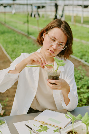 Scientist in greenhouse inspecting plant seedling while taking notes with clipboard on table, illustrating precision in agricultural researchの写真素材