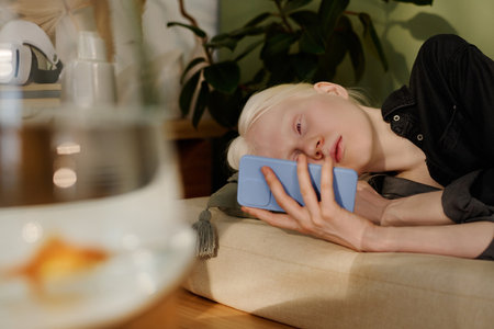 Gen Z girl with albinism scrolling social media news feed on smartphone while relaxing on couch in living roomの写真素材