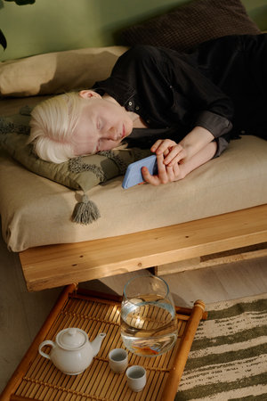 Vertical high angle view of young Caucasian girl with albinism surfing Internet on smartphone while relaxing on couch in living room in morningの写真素材