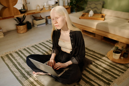 Gen Z female writer with albinism sitting on floor in cozy living room and working on laptop in morningの写真素材