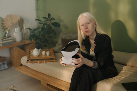 Young Caucasian woman with albinism holding VR headset in hands sitting on couch in living room and...