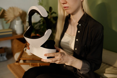 Medium section shot of young woman with long white hair holding modern VR headset in her hands while sitting on couch at homeの写真素材