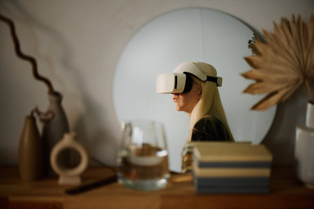 Round mirror with young white-haired woman wearing VR headset reflected in it on wooden shelf decorated with books and vesselsの写真素材