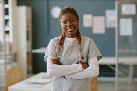 Portrait of smiling African American nurse with arms crossed, wearing uniform in medical facility background, convey professional confidence and warmth while standingの写真素材